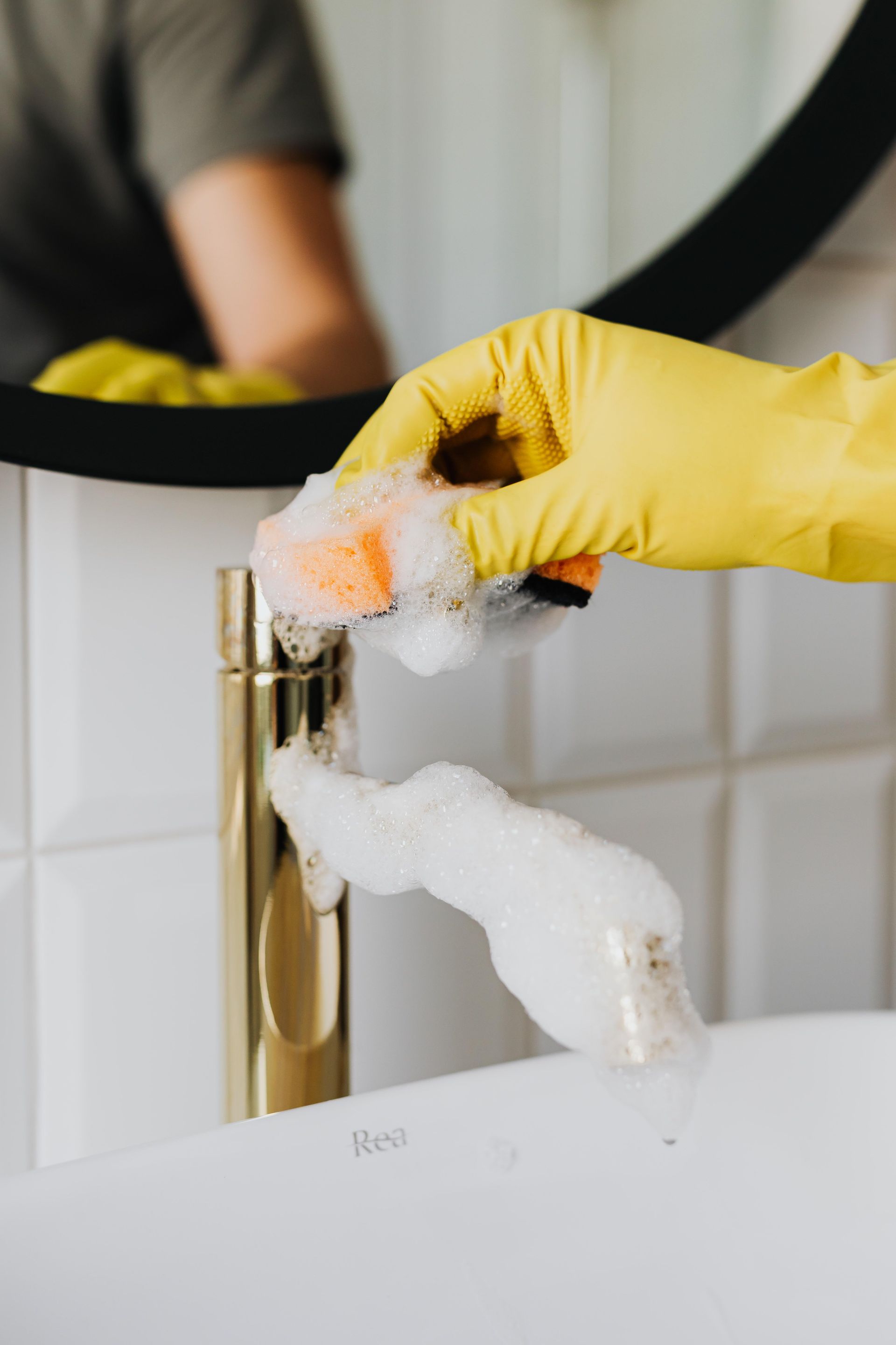 Anonymous person washing golden shiny faucet and sink in bathroom while wearing yellow latex glove during household care routine work